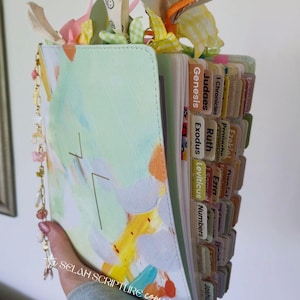 May include: A light green Bible with colorful abstract art on the cover. The Bible has labeled tabs and decorative embellishments at the top. The text "SELAH SCRIPTURE company" is visible.