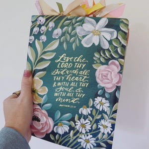 May include: A rectangular book with a dark green cover featuring hand-painted white, pink, and yellow flowers. The cover displays gold cursive text that reads: Love the LORD THY God with all THY heart & WITH ALL THY Soul & WITH ALL THY mind MATTHEW 22:37.