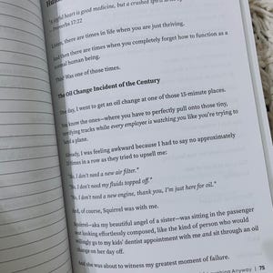 May include: Open book with text. The page reads "Day 18: When You Completely Forget How to Human." The text describes an oil change experience. The book is open to page 75, with the title "Holy Shenanigans: Grace, Grief & Laughing Anyway."