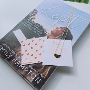 May include: A gold-toned necklace with a semi-circular pendant featuring the word Kept, resting on a book titled Kept by Emily Hampton, alongside two small square cards, one with an orange polka dot pattern and one with printed text.