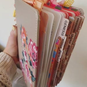 May include: A beige Bible with a floral design in pink, orange, and green. The pages are tabbed with the names of the books of the Bible. Pink and peach ribbons are tied at the top.