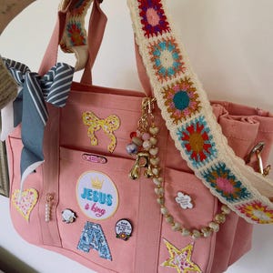 May include: Pink tote bag with a colorful crochet strap. The bag features multiple patches, including a bow, star, and the text "JESUS & king." A charm with the letter "A" and a beaded strand hang from the bag.
