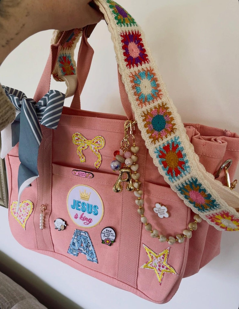 May include: Pink tote bag with a colorful crochet strap. The bag features multiple patches, including a bow, star, and the text "JESUS & king." A charm with the letter "A" and a beaded strand hang from the bag.