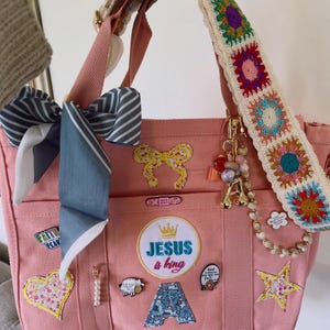 May include: Pink canvas tote bag with multiple pockets, decorated with patches, pins, and a bow. Features a crocheted strap with colorful squares and a gold charm. The text "JESUS is king" is visible on the front.