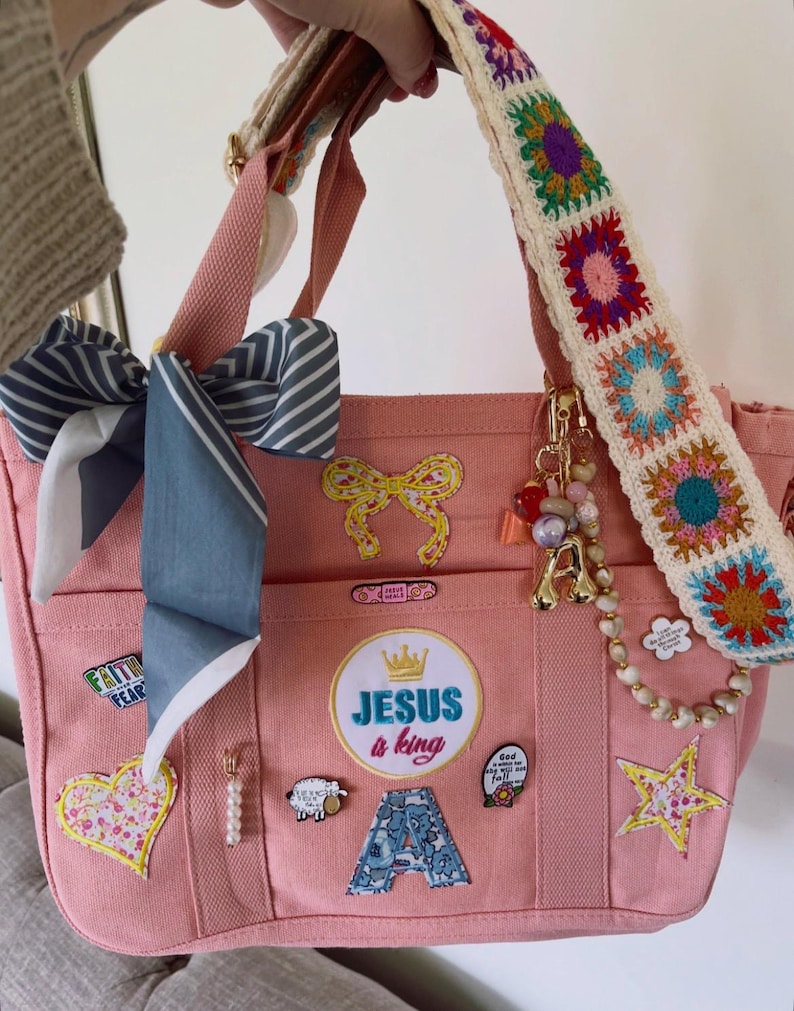 May include: Pink canvas tote bag with multiple pockets, decorated with patches, pins, and a bow. Features a crocheted strap with colorful squares and a gold charm. The text "JESUS is king" is visible on the front.