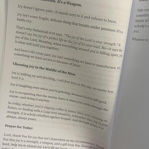 May include: Open book with text about joy, including the phrases "Joy is Not an Emotion. It's a Weapon" and "Choosing Joy in the Middle of the Mess." The pages are white with black text.