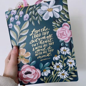 May include: A dark teal book cover with floral illustrations and gold lettering. The cover features pink roses, white daisies, and other flowers. The text reads "Love the LORD THY God with all THY heart & WITH ALL THY Soul & WITH ALL THY mind" from Matthew 22:37.