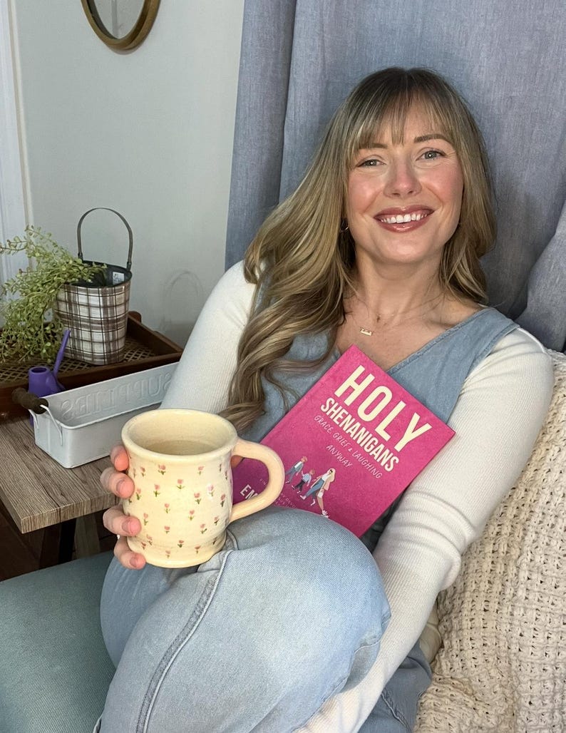 May include: A cream-colored ceramic mug with a floral pattern is held near a pink book titled "Holy Shenanigans." The book's cover features illustrations and text. The person is wearing a white long-sleeve shirt and blue denim overalls.