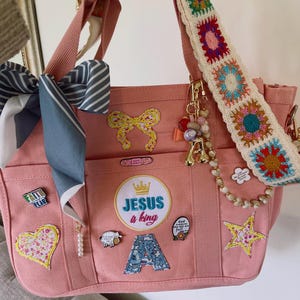 May include: Pink canvas tote bag with multiple pockets, adorned with patches, pins, and a crocheted strap. Features a bow, star, and the text "Jesus a King". Includes a decorative scarf and beaded charms.