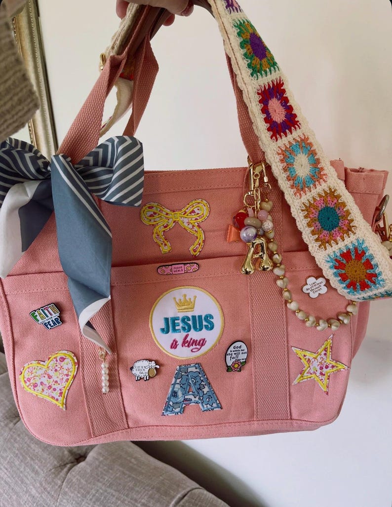 May include: Pink canvas tote bag with multiple pockets, adorned with patches, pins, and a crocheted strap. Features a bow, star, and the text "Jesus a King". Includes a decorative scarf and beaded charms.
