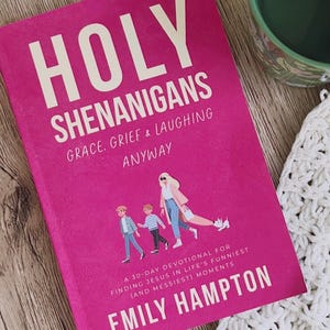 May include: A hot pink book titled "HOLY SHENANIGANS" with white text. The book cover features the text "GRACE, GRIEF & LAUGHING ANYWAY" and "A 30-DAY DEVOTIONAL FOR FINDING JESUS IN LIFE'S FUNNIEST (AND MESSIEST) MOMENTS" by Emily Hampton. A green mug is in the background.