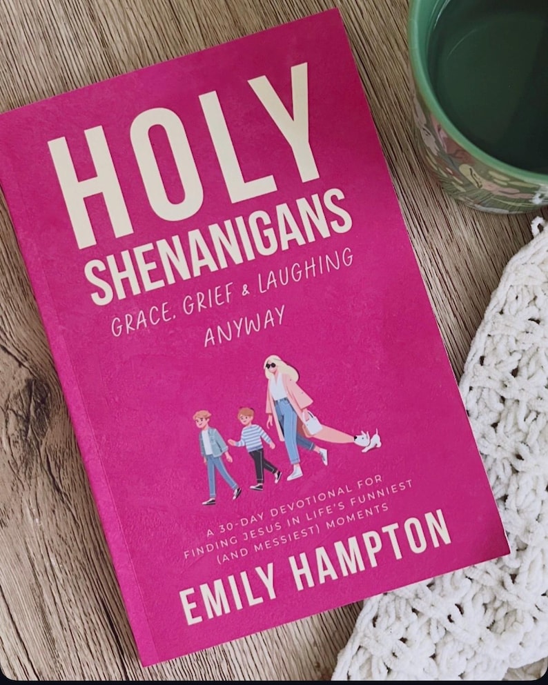 May include: A hot pink book titled "HOLY SHENANIGANS" with white text. The book cover features the text "GRACE, GRIEF & LAUGHING ANYWAY" and "A 30-DAY DEVOTIONAL FOR FINDING JESUS IN LIFE'S FUNNIEST (AND MESSIEST) MOMENTS" by Emily Hampton. A green mug is in the background.