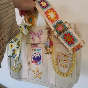 May include: Cream-colored tote bag with a colorful crochet strap. The bag features patches with text and floral designs, a yellow beaded chain, and a gold-toned charm. The strap is adorned with crochet squares in various colors.