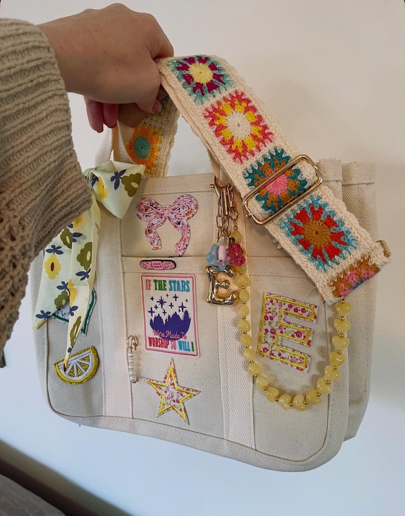 May include: Cream-colored tote bag with a colorful crochet strap. The bag features patches with text and floral designs, a yellow beaded chain, and a gold-toned charm. The strap is adorned with crochet squares in various colors.