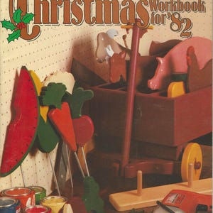 May include: The cover of a vintage "Make-it-Yourself Christmas Workbook for '82" is displayed. The image features wooden Christmas crafts, including a pink pig in a wagon, and colorful wooden food cutouts. Paint cans and a drill are also visible.