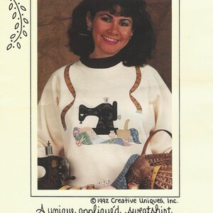 May include: A cream-colored sweatshirt with a black collar and a sewing machine applique. The design includes a measuring tape and the text "STITCHES N' STUFF". The sweatshirt features handmade porcelain buttons. The image includes the text "Creative Uniques, Inc."