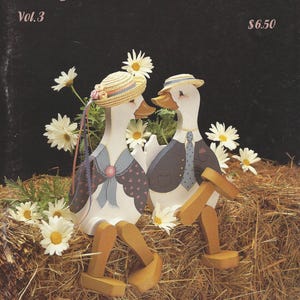 May include: The cover of "Keepsake Sampler Vol. 3" features two painted wooden ducks, adorned with hats and clothing, sitting on a bed of hay with daisies. The ducks are painted white with brown beaks and wooden legs. The text "Keepsake Sampler" is at the top.