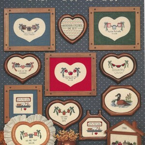 May include: A collection of framed cross-stitch samplers with heart, house, and oval shapes. The samplers feature various designs, including houses, hearts, and text like "Welcome to Our Neighborhoods" and "From Bow to Bow". The color palette includes red, blue, green, and brown.