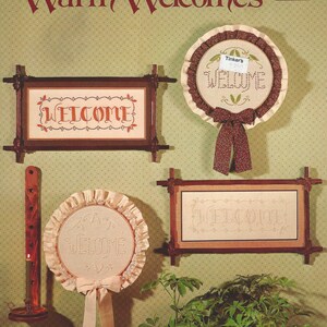 May include: Image of cross-stitch "Welcome" signs in wooden frames, with bows and decorative trim. The background is light green with a polka dot pattern. The text "Warm Welcomes" is displayed at the top. The signs are in a vintage style.