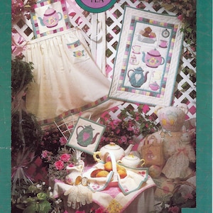 May include: A tea party-themed image with a white apron, a quilted wall hanging, and a table setting. The apron and wall hanging feature teapot designs. The text "TIME FOR TEA" is in a circle at the top. The text "MUMM'S THE WORD" is at the bottom.