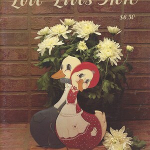 May include: A wooden duck couple figurine with a red and white polka-dotted dress and a blue overall. The figurine is in front of a brick wall and a bouquet of white flowers. The text "Love Lives Here" is at the top.