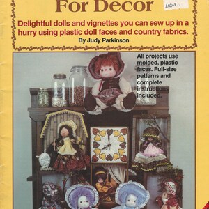 May include: A book cover titled "CALICO DOLLS For Decor" with various dolls and vignettes displayed. The cover text mentions using plastic doll faces and country fabrics. The image includes a shelf with several dolls, a clock, and jars.