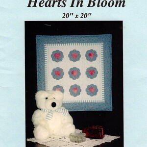 Peut inclure: Une courtepointe avec une bordure bleue et un centre blanc avec neuf motifs en forme de cœur. La courtepointe mesure 50,8 cm x 50,8 cm. L'image comprend également un ours en peluche blanc, une nappe en dentelle et le texte "Paper Pieces" et "Hearts In Bloom."