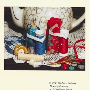 May include: Image of a needlecase and chatelaine doll. The image features a white teapot with gold floral designs in the background. In the foreground are spools of thread, thimbles, and small dolls. The text "NEEDLECASE CHATELAINE DOLL" is at the top.