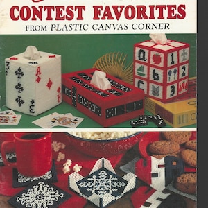 May include: A book cover featuring various plastic canvas projects. The title reads "Creative Contest Favorites" with a subtitle "From Plastic Canvas Corner." The image showcases tissue box covers, coasters, and other craft items.