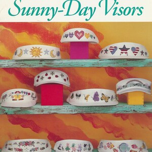 May include: A collection of cross-stitch visors featuring embroidered patterns like hearts, stars, and butterflies. The white visors are displayed on shelves. The image includes the text "Sunny-Day Visors".