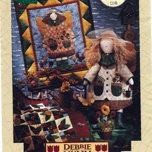 Könnte beinhalten: Farbenfrohes Bild einer handgefertigten Puppe und eines Quilts mit Engelsmotiv. Die Puppe trägt einen Hut und einen Mantel und hält einen Korb. Der Quilt zeigt ein Engeldesign mit Sonnenblumen. Text enthält "Greta, The Garden Angel" und "Debbie Mumm Friendship Club".