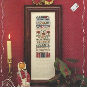 Könnte beinhalten: Ein gerahmter Sampler mit Kreuzstich-Alphabet und floralen Designs. Der Text "THE PRACTICAL SAMPLER" steht oben. Eine Kerze, eine Puppe und eine Pflanze befinden sich im Vordergrund. Das Preisschild lautet 5,00 $.