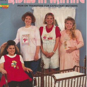 May include: A craft pattern book titled Ladies-In-Waiting Iron-On Transfers For Expectant Mothers by Marina Anderson. The cover features four individuals wearing clothing decorated with iron-on transfers, positioned near a wooden baby cradle.