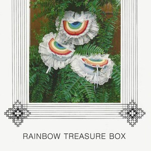 Peut inclure: Image d'un patron d'artisanat intitulé "Rainbow Treasure Box". L'image présente trois ornements en forme d'arc-en-ciel avec des bords à volants et une garniture en dentelle, sur fond de feuillage vert. Le patron est de Cross 'N Patch, Box 132, Millville, Utah 84326.