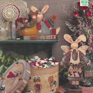 Op de afbeelding: Een vintage cover van "Makin' Pretties" met handgemaakte konijnenbeeldjes, een doos vol knopen en een decoratieve hoed. De cover bevat de tekst "Makin' Pretties" en "By Michele Deaton".