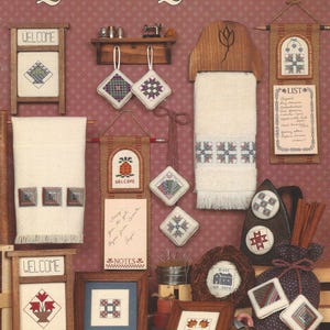 May include: A collection of cross-stitch projects, including framed pieces, welcome signs, and hanging ornaments. The image features various geometric and floral designs in a rustic setting. The text "Quilted Quickies" is visible.