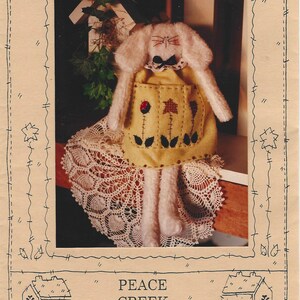 Könnte beinhalten: Handgemachte Blossom Bunny-Puppe, ca. 48 cm groß, mit abnehmbarem Knopfkopf. Die Puppe hat einen cremefarbenen Körper, eine gelbe Schürze mit floralen Stickereien und wird auf einer Spitzendecke präsentiert. Der Text "Peace Creek Collections" ist ebenfalls sichtbar.