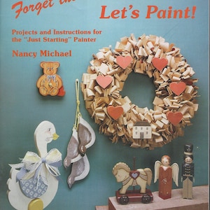 May include: A book cover titled "Forget the Dust - Let's Paint!" featuring painted wooden crafts. A wreath with red hearts, a goose, a teddy bear, and other decorative items are displayed. The book is a Jackie Shaw Studio Publication.