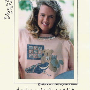 May include: A pale pink sweatshirt with an appliqued design of a teddy bear and a cat. The design includes a blue and white quilt square and a blue patterned blanket. The text "QUILT'N PALS" is at the top.