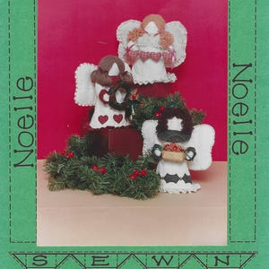 May include: Three fabric angel dolls with yarn hair, white dresses, and wings, arranged with a green pine garland featuring red berries. The green border frame includes the text Noelle and the phrase SEWN IN LOVE with heart icons.