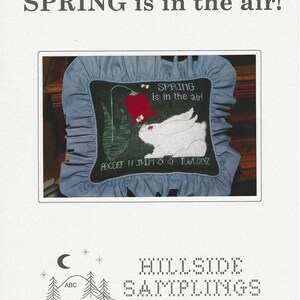 May include: A cross-stitch pattern featuring a white rabbit and a red flower, with the text "SPRING is in the air!". The design is on a black background, framed by a blue and white striped fabric. The pattern is from Hillside Samplings.