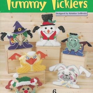 May include: A craft kit cover featuring six seasonal plastic canvas characters: a witch, snowman, Frankenstein's monster, duck, bunny, and elf. The title "Tummy Ticklers" is displayed in yellow, with the text "Plastic Canvas" above.