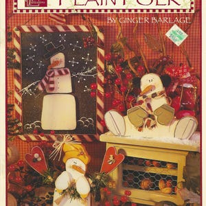 May include: A magazine cover featuring the title "PLAIN FOLK" and various wooden snowman decorations. The snowmen are painted with winter attire and accessories. The background includes red berries and a checkered pattern.