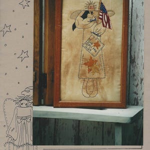 May include: A framed Americana angel stitchery. The angel holds a US flag and a sign that reads "God Bless the USA." The artwork is 9" x 12" and features a rustic, folk art style with a brown wooden frame. The angel is wearing a dress with autumn leaves.