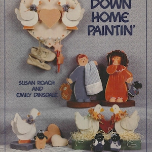 Puede incluir: Una colección de artesanías de madera pintada, que incluye gansos, adornos de pared en forma de corazón y figuras de niños. La imagen también presenta el texto "DOWN HOME PAINTIN'" y los nombres "SUSAN ROACH AND EMILY DINSDALE".