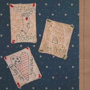 Op de afbeelding: Handgestikte textielkunst met drie rechthoekige patches op een blauwe achtergrond met witte sterren. De patches tonen geborduurde ontwerpen van een kous, een bijenkorf en een hart met tekst. De woorden "Old Wool Stocking", "Beeskep" en "Joy Can See" zijn zichtbaar.