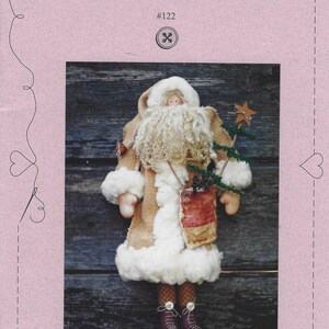 May include: A craft pattern cover titled Sweet Stitches Country Kringle #122 on a light pink background with a dashed border and heart icons. The central image shows a fabric Santa doll with a white beard, tan coat, and a small tree.