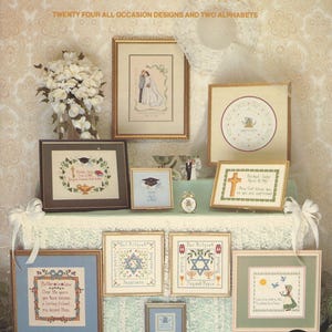 Könnte beinhalten: Eine Präsentation von gerahmten Kreuzstich-Samplern mit verschiedenen Designs, darunter Hochzeits-, Abschluss- und religiöse Themen. Der Text "Loving Memories" steht oben, zusammen mit dem Namen des Designers. Die Farbpalette umfasst Gold, Blau, Grün und Braun.