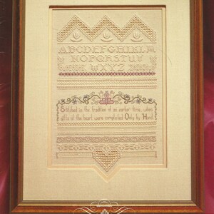 May include: Framed cross-stitch sampler with a beige background and a gold-colored wooden frame. The sampler features embroidered letters, decorative borders, and the text "Stitched in the tradition of an earlier time, when gifts of the heart were completed Only by Hand."