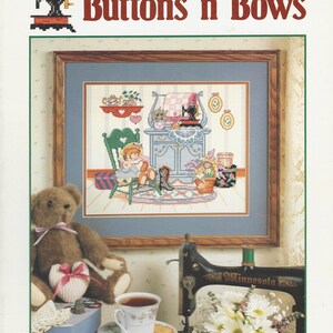 May include: A craft pattern titled "Buttons 'n Bows" by Barbara Mock. The image features a framed cross-stitch scene with a teddy bear, sewing machine, and teacup. A brown teddy bear with a pink ribbon sits next to a teacup and saucer.
